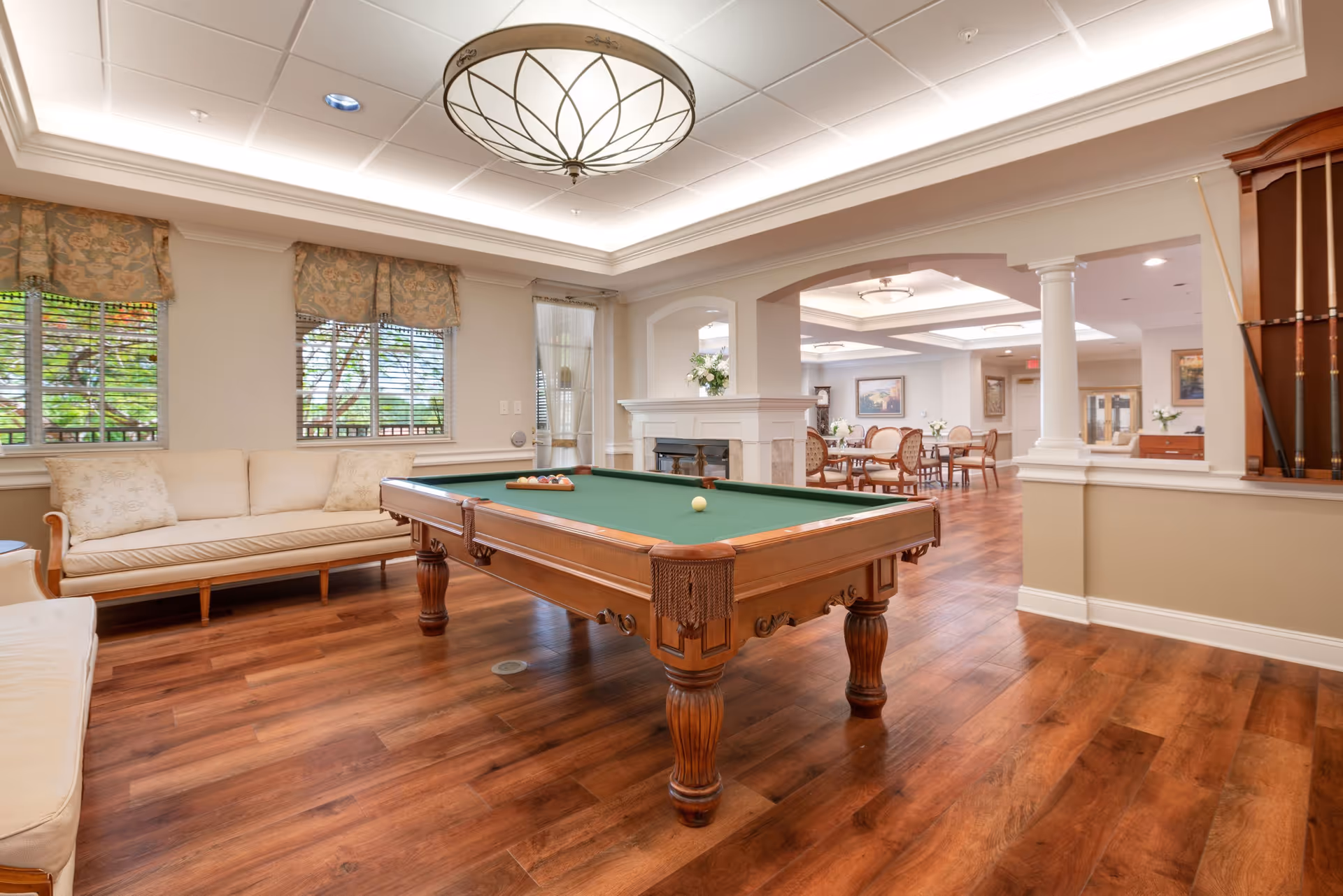A spacious senior living common area featuring a pool table in the center with a green felt surface. Surrounding the pool table are cushioned benches and chairs. The room has large windows with floral valances, wooden flooring, and a decorative ceiling light fixture. In the background, there is a dining area with tables and chairs, separated by white columns and a half wall with a fireplace.