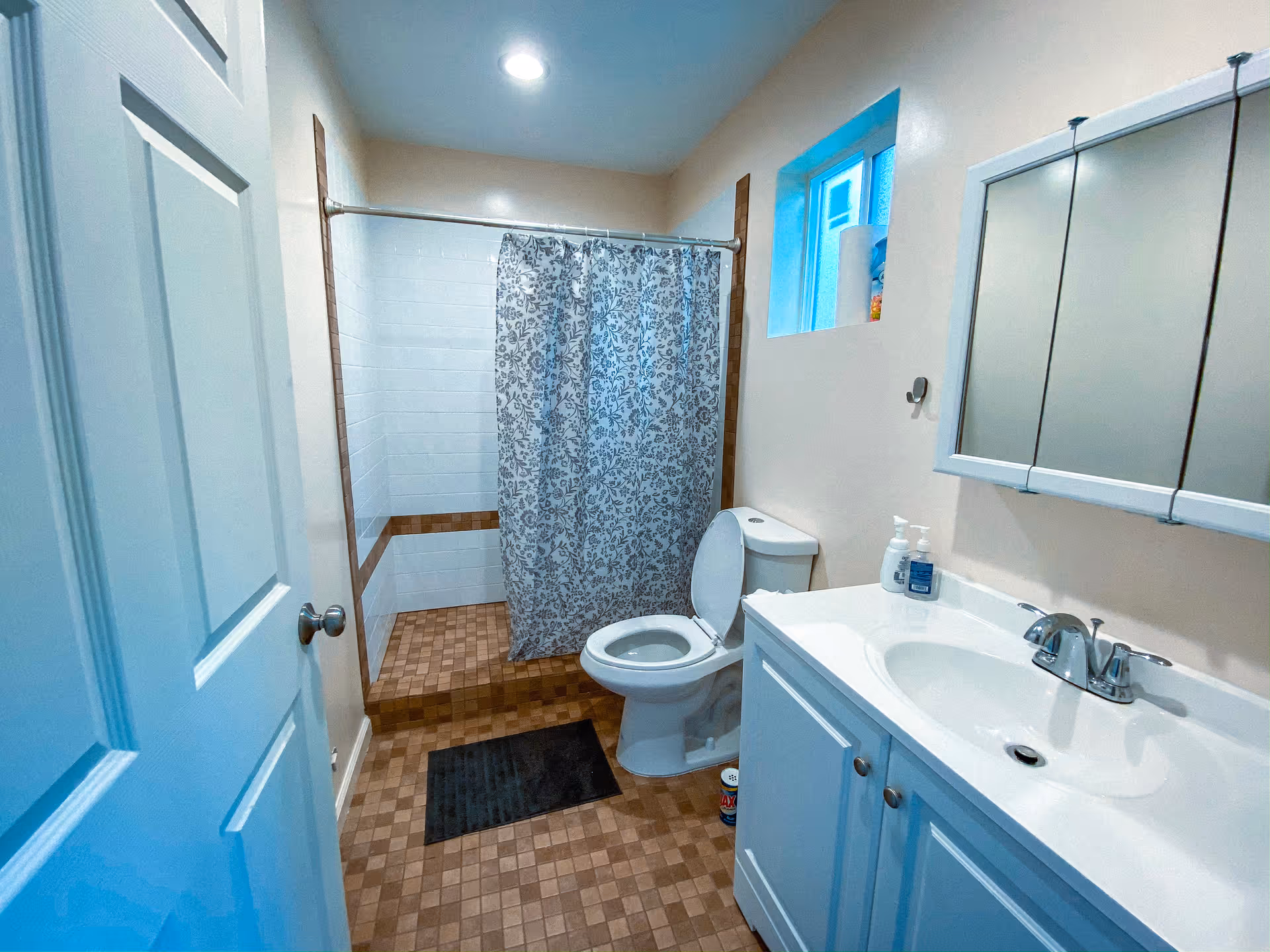 A bathroom with a white door partially open, a toilet with the lid up, a white sink with a faucet and soap dispensers, a mirrored medicine cabinet above the sink, a small window, and a shower area with a floral-patterned shower curtain and tiled walls and floor.