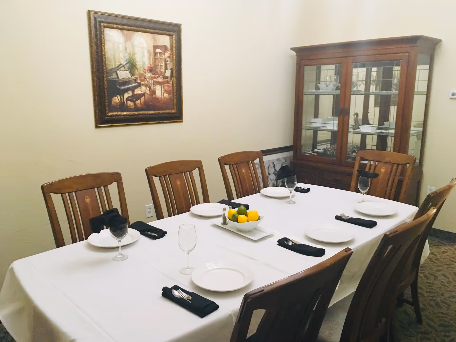 A dining room with a rectangular table covered with a white tablecloth, set with white plates, black napkins, and wine glasses. There are eight wooden chairs around the table. A bowl of lemons and limes is placed in the center of the table. On the wall, there is a framed painting depicting a piano and a cozy room. A wooden cabinet with glass doors containing dishes is positioned against the wall.