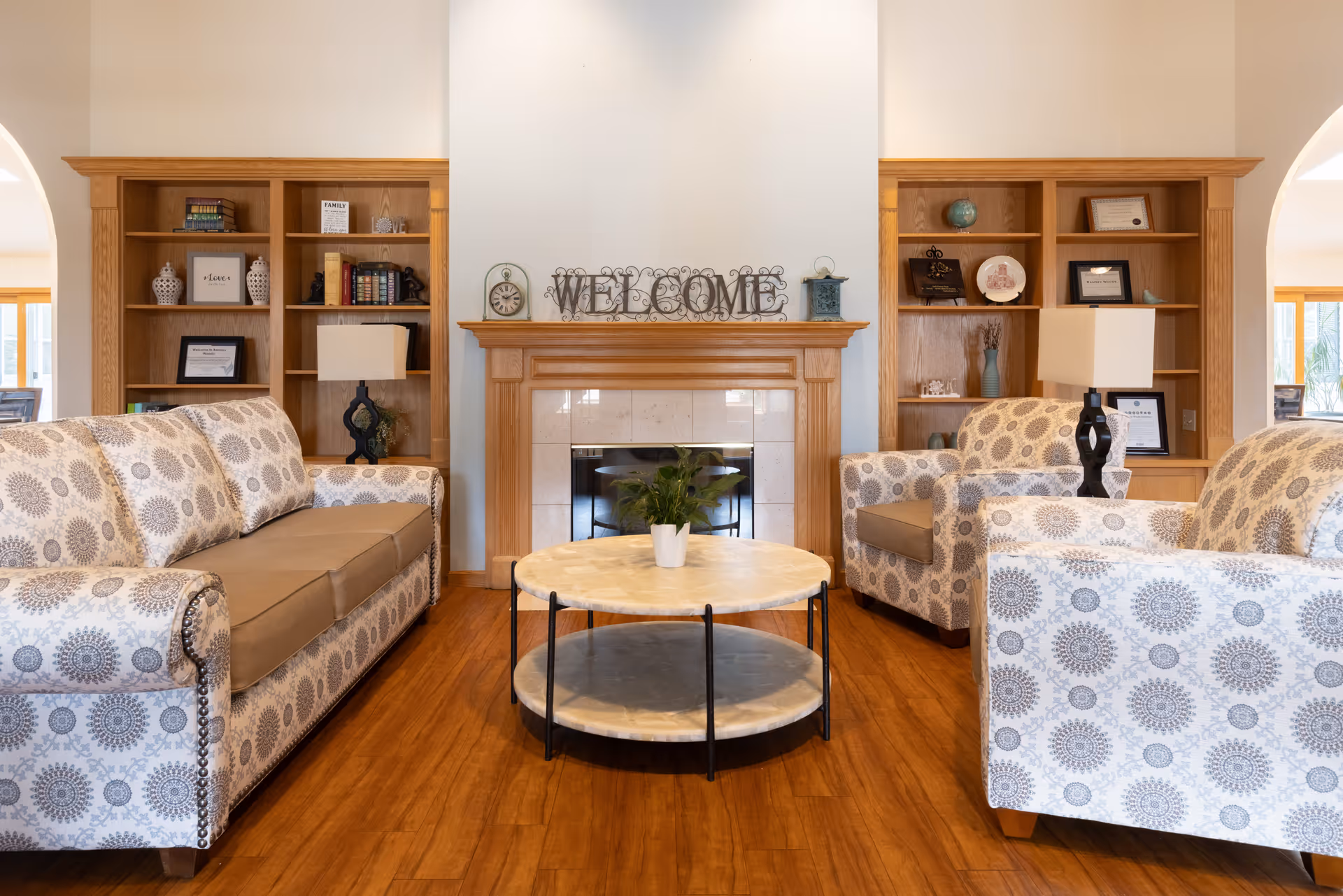 A cozy living room with patterned sofas and armchairs arranged around a round marble coffee table. Behind the seating area is a wooden fireplace mantel with a decorative 'WELCOME' sign on top. Flanking the fireplace are built-in wooden bookshelves filled with books, framed pictures, and decorative items. The room has warm wooden flooring and soft natural lighting.