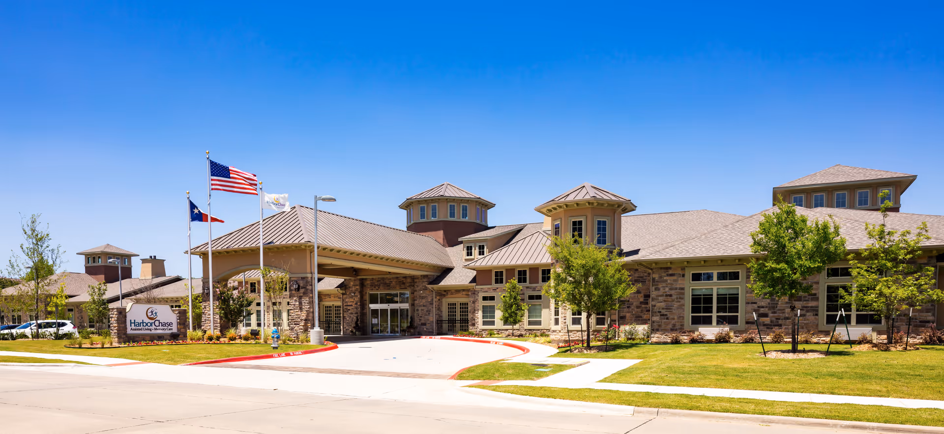 Exterior view of a senior living facility building under a clear blue sky. The building has a stone facade with multiple peaked roofs and large windows. There are three flagpoles in front displaying the American flag, the Texas state flag, and another flag. The facility sign reads 'HarborChase'. The surrounding area includes a well-maintained lawn, small trees, and a paved driveway.