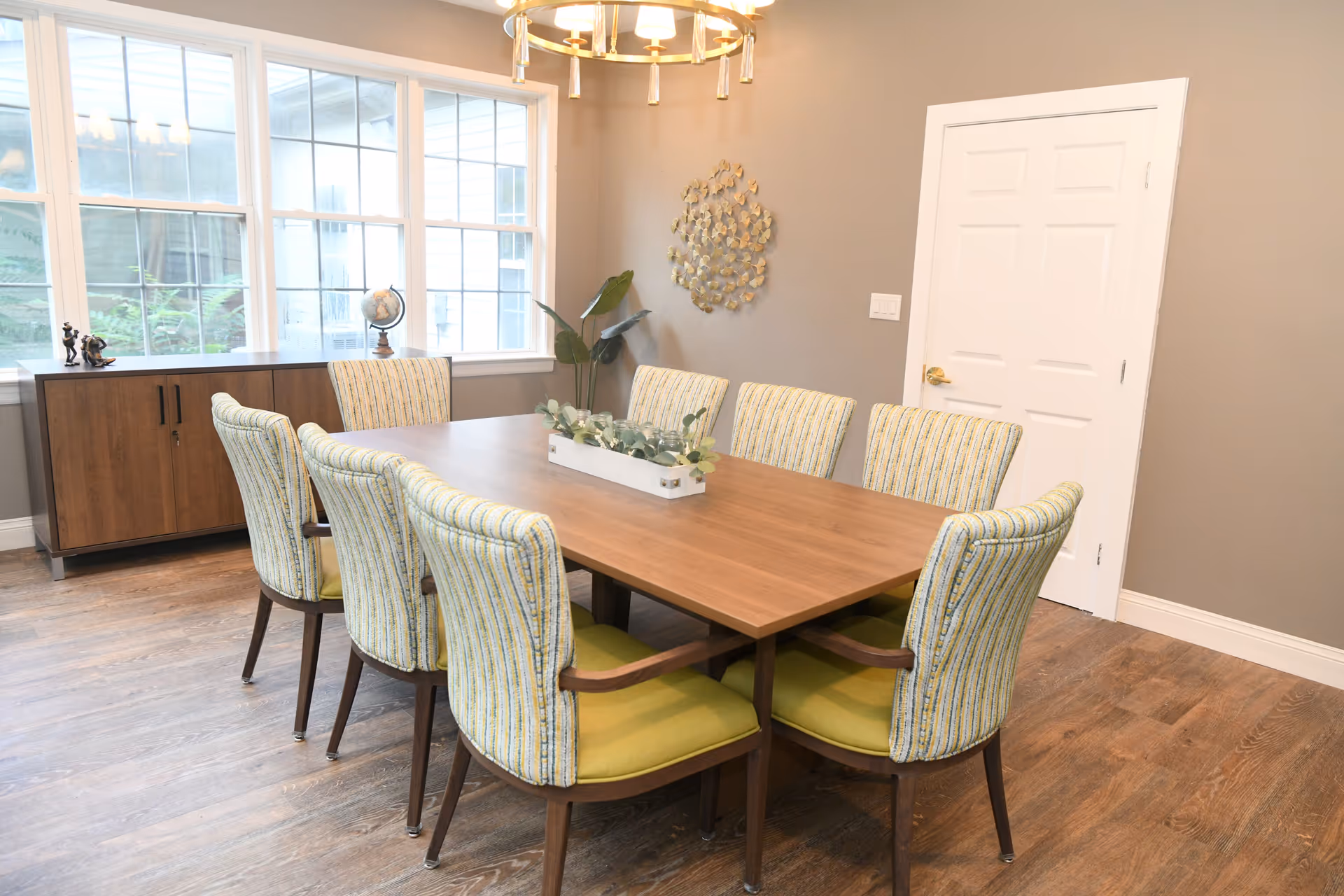 Bright dining room with a rectangular wooden table surrounded by eight upholstered chairs, a sideboard under large windows, and wall decor.