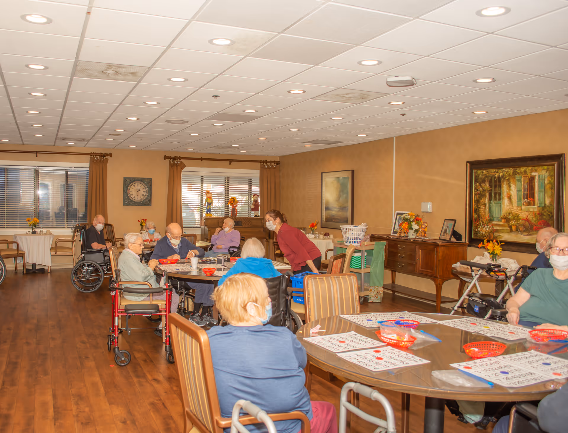 Elderly residents wearing masks sit around tables in a well-lit communal activity/dining room with bingo cards while staff assist.