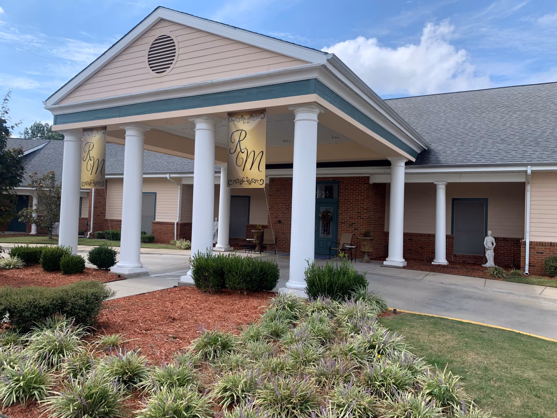 Front exterior view of a building with a covered entrance supported by white columns. The building has beige siding with brick accents and a gray shingled roof. There are two hanging banners with the letters 'RM' on the covered entrance. Landscaping with bushes and mulch is visible in the foreground under a partly cloudy sky.