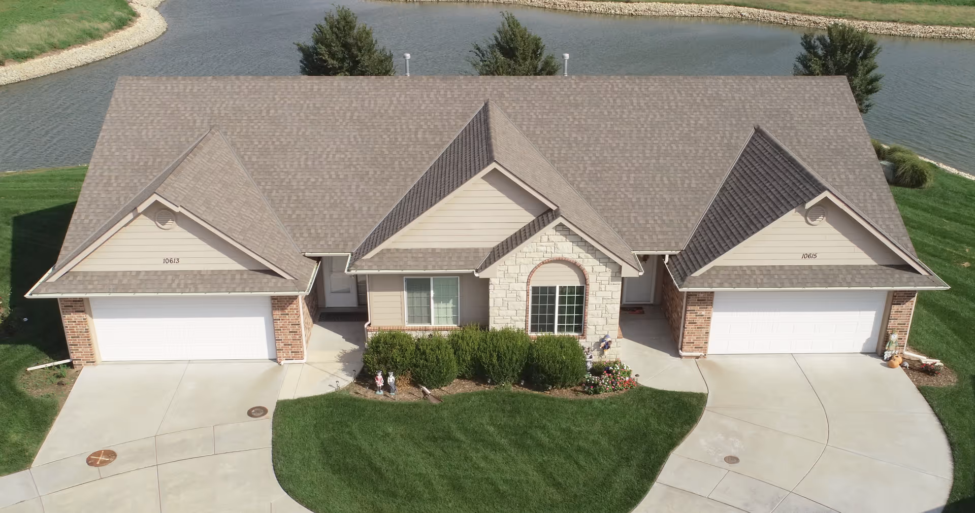 Aerial view of a duplex-style residential building with two garages, beige siding, brick and stone accents, and a well-maintained lawn in front. The building is situated near a body of water with trees along the shoreline.