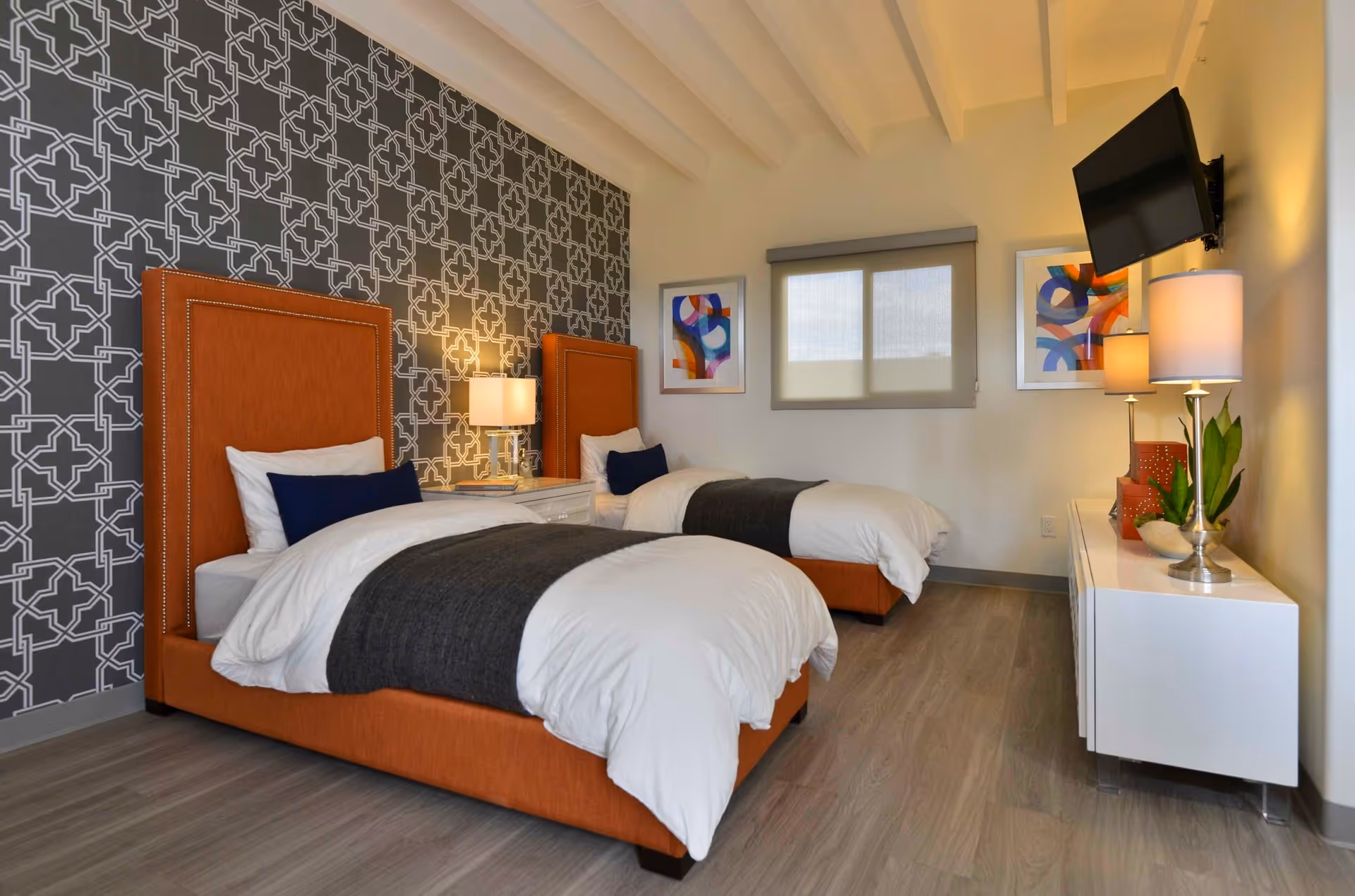 Two twin beds with orange headboards in a modern, well-lit bedroom featuring a patterned accent wall, bedside lamps, and a wall-mounted TV.
