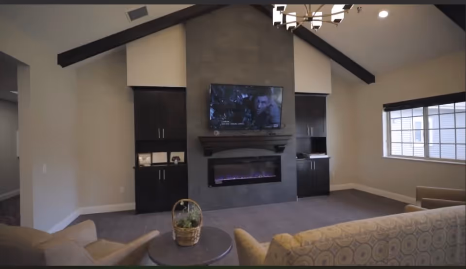 A cozy living room with a modern fireplace set in a dark stone wall, flanked by two black cabinets. Above the fireplace is a flat-screen TV displaying a scene from a movie. The room has a high ceiling with exposed dark wooden beams and a chandelier. There is a window with blinds on the right wall, and comfortable seating including an armchair and a patterned sofa facing the fireplace. A small round table with a basket of plants is in the center.