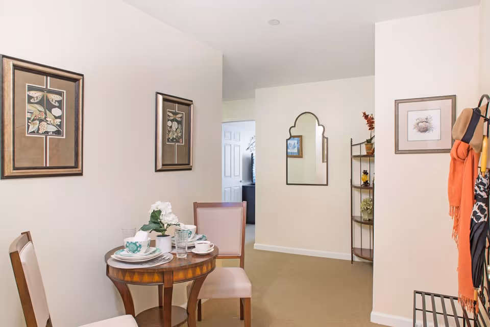 A small dining area with a round wooden table set for two with plates, cups, and a flower centerpiece. Two chairs with light upholstery are placed around the table. The walls are decorated with framed botanical prints and a decorative mirror. A corner shelf with plants and decorative items is visible, along with a coat rack holding scarves and umbrellas.
