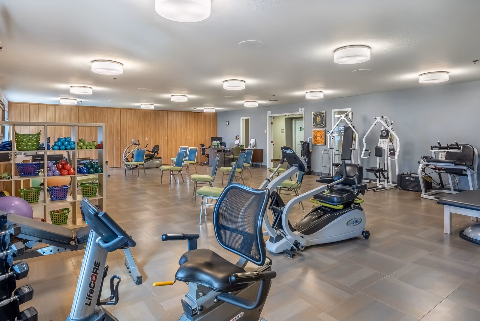A spacious fitness room in a senior living facility with various exercise equipment including stationary bikes, weight machines, and a rack with colorful exercise balls and baskets. Several chairs are arranged around the room, and the walls are light-colored with wood paneling on one side.