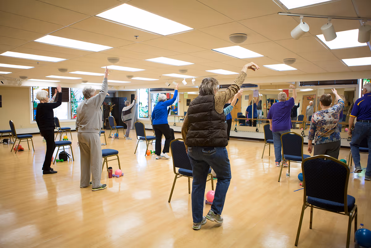 A group of elderly people participating in a seated exercise class in a spacious room with wooden floors, chairs spaced apart, and large mirrors on one wall. They are raising one arm while standing or sitting, engaging in a coordinated movement.