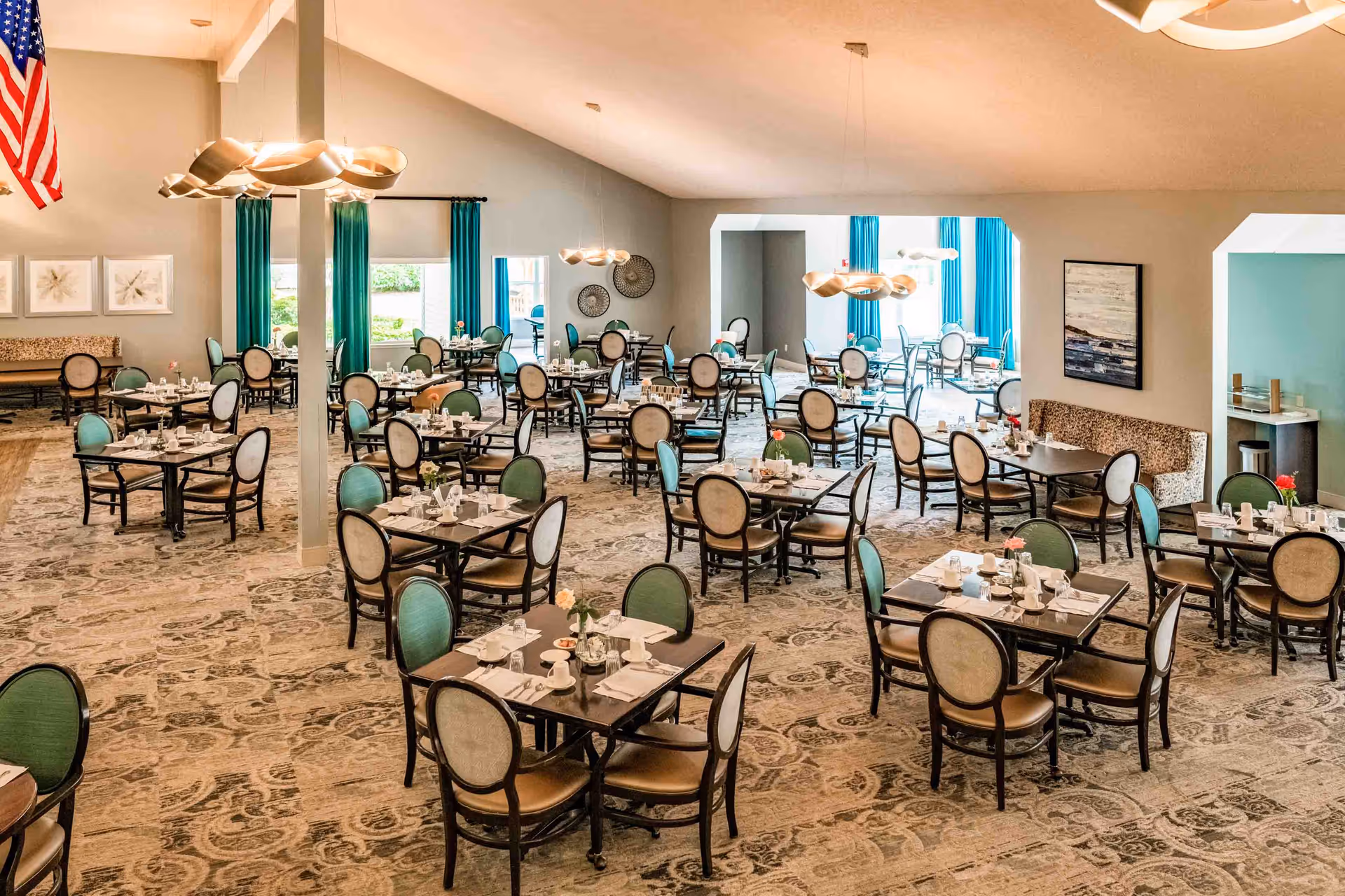 A spacious dining room with multiple tables and chairs arranged neatly. Each table is set with white napkins, cups, and small flower vases. The room features large windows with teal curtains, modern light fixtures, and a patterned carpet. An American flag is visible on the left side of the room.