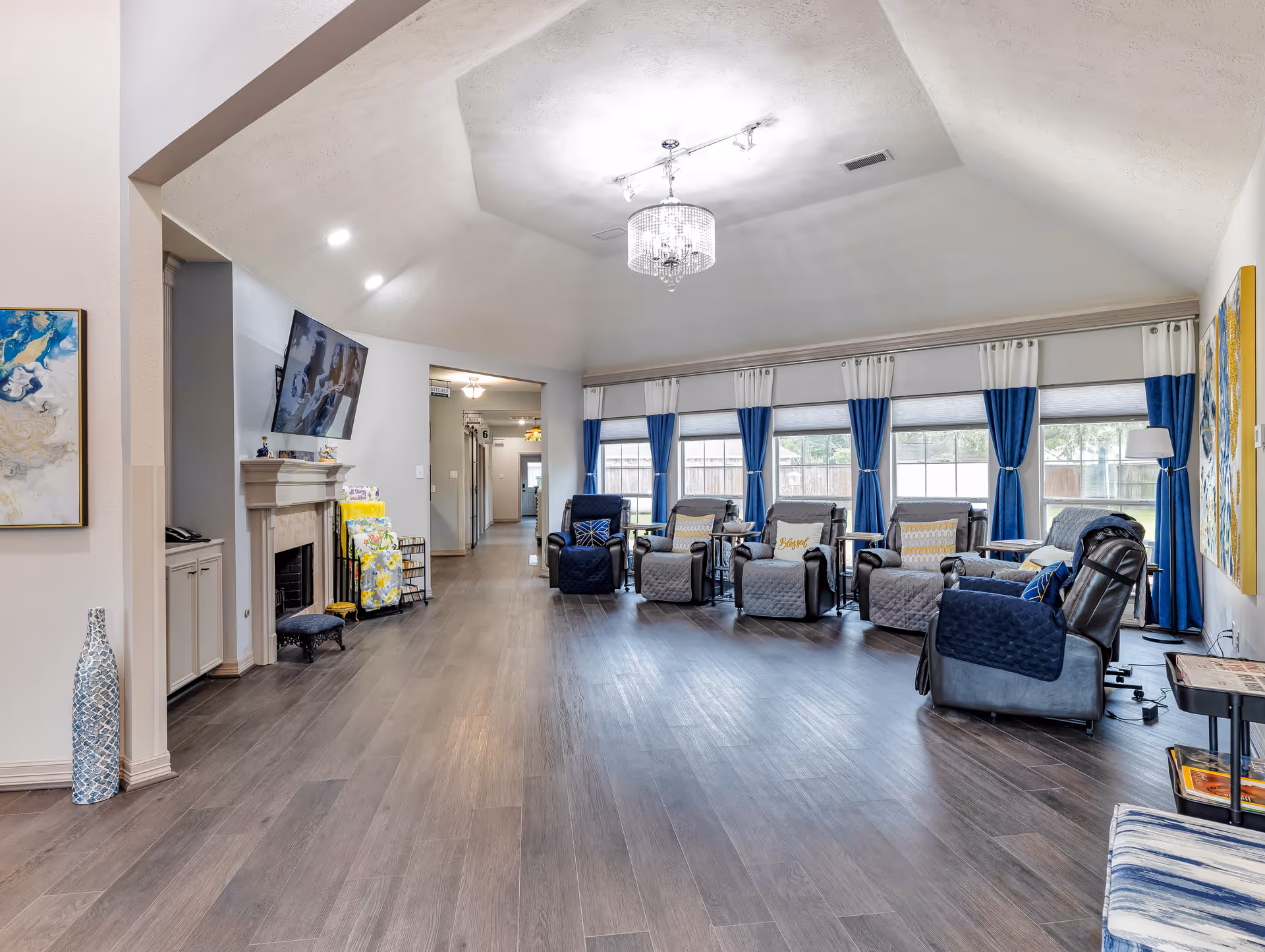 Spacious communal living room with a row of recliners facing large windows with blue curtains, a chandelier, and a TV above a fireplace.