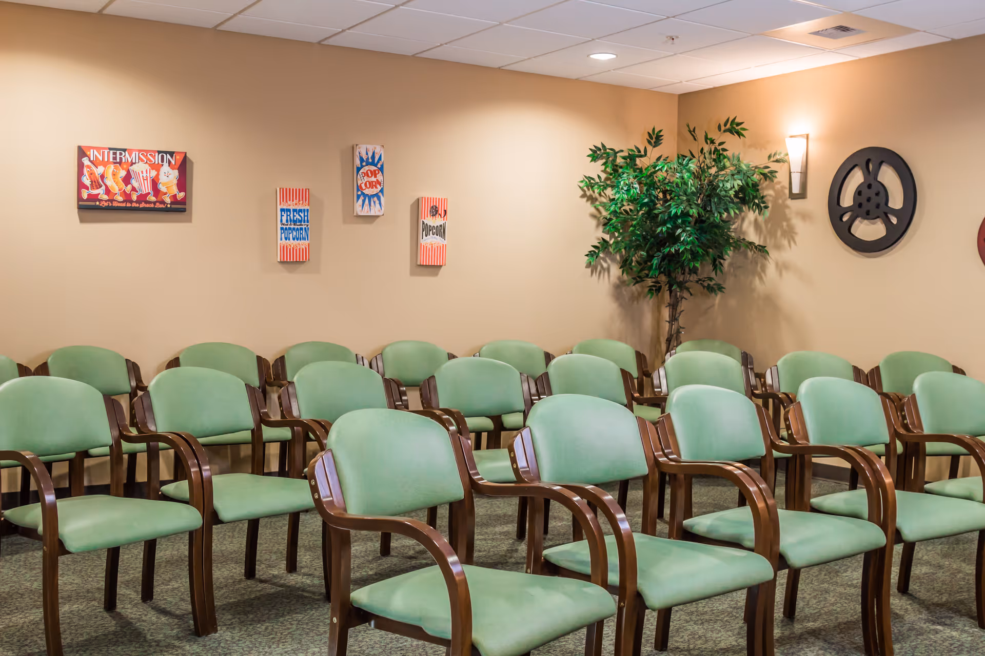 Rows of green upholstered chairs arranged in a small screening/activity room with movie-themed wall decor and a potted plant.