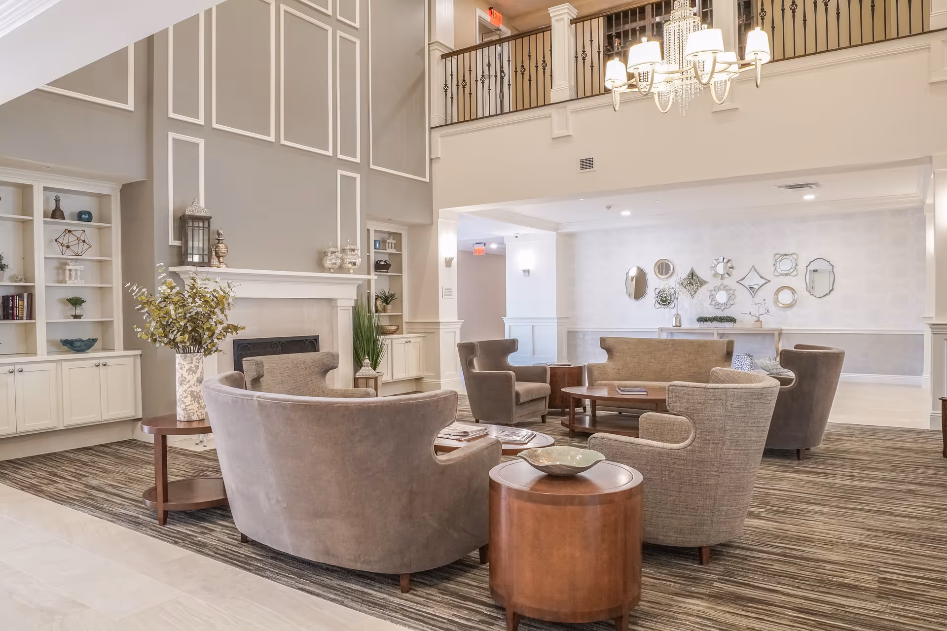 A spacious and elegant living room area in a senior living facility with a high ceiling, a fireplace, and a chandelier. The room features several comfortable armchairs and sofas arranged around wooden coffee tables. Decorative mirrors and plants adorn the walls and shelves, creating a warm and inviting atmosphere.