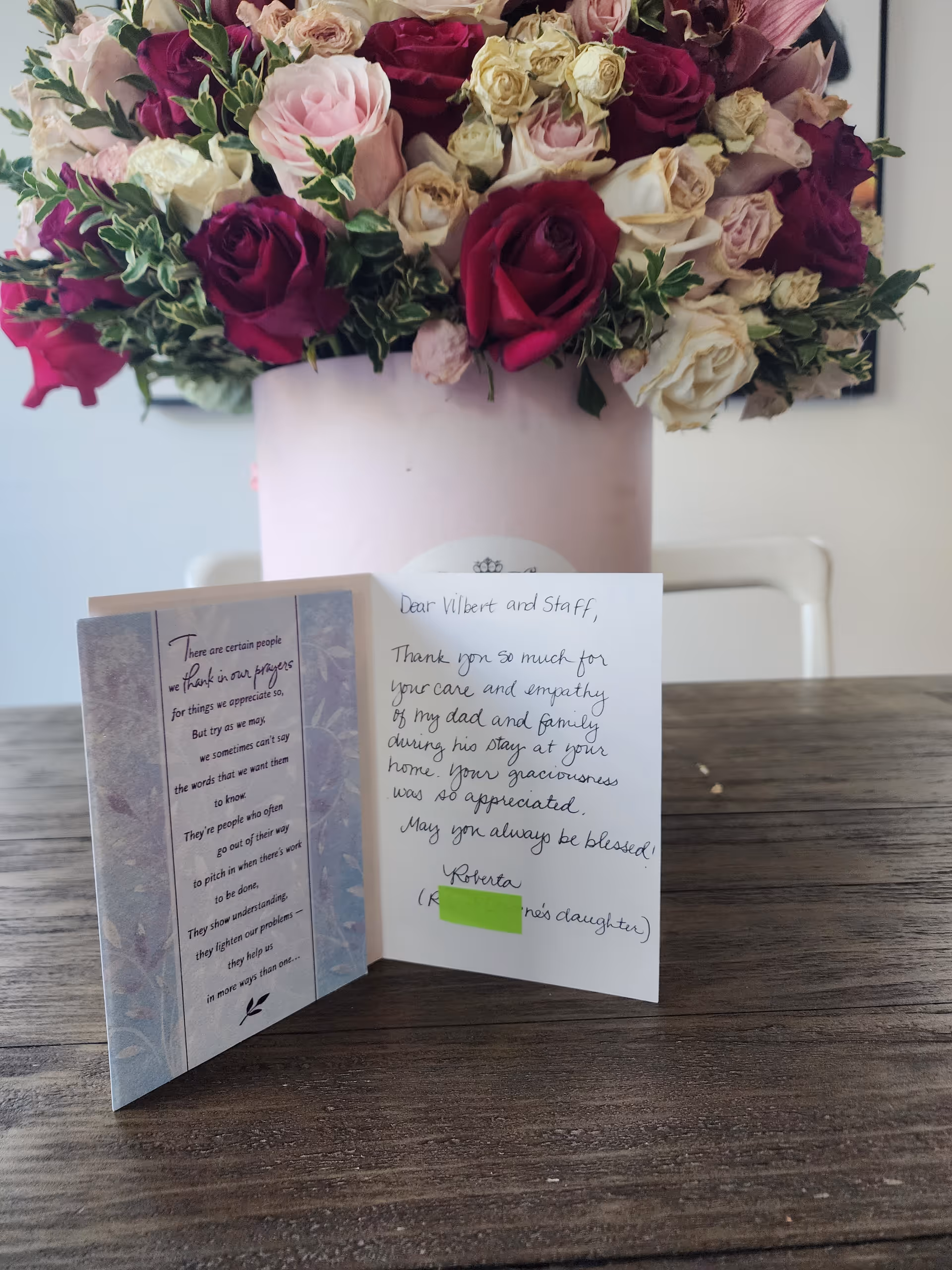 A bouquet of mixed roses in shades of red, pink, and cream in a light pink vase on a wooden table. In front of the vase is an open thank-you card with a handwritten message expressing gratitude to Vilbert and staff for their care and empathy during a family member's stay.
