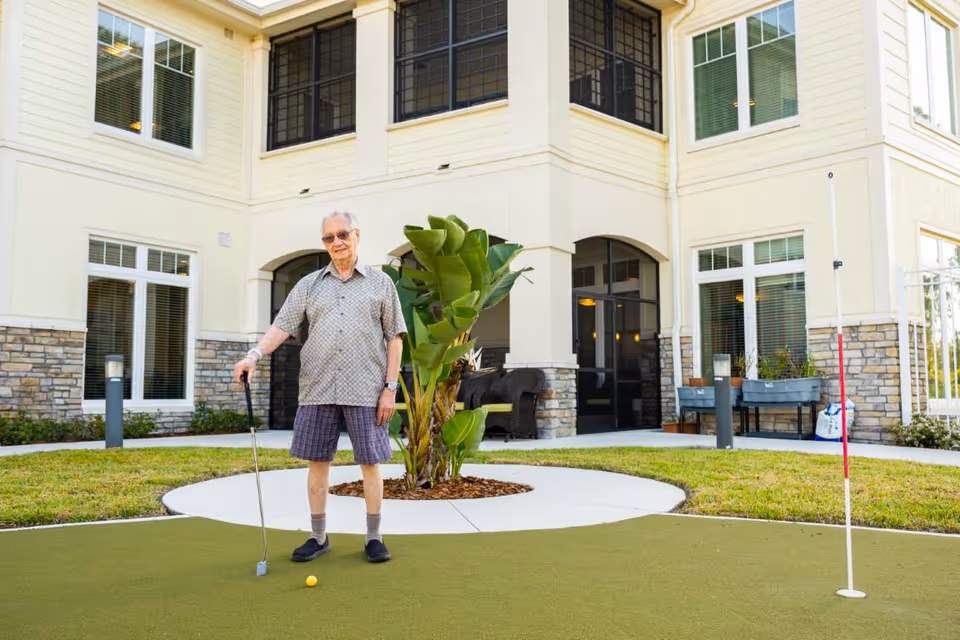 An elderly man standing on a putting green outside a senior living facility, holding a golf putter and smiling. The building behind him has large windows and a patio area with outdoor furniture and plants.
