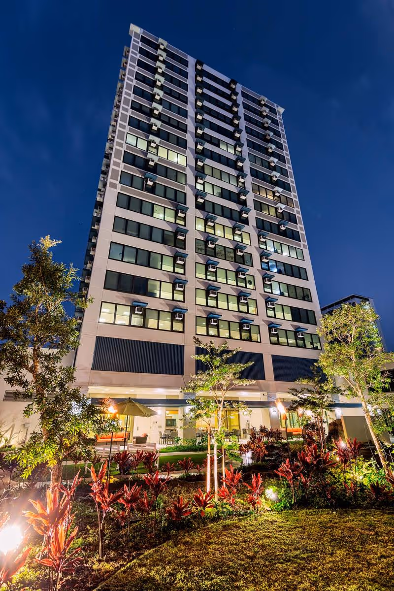 Tall multi-story building at night with many windows lit up, surrounded by landscaped garden with trees and plants, and outdoor seating area with tables and umbrellas at the ground level.