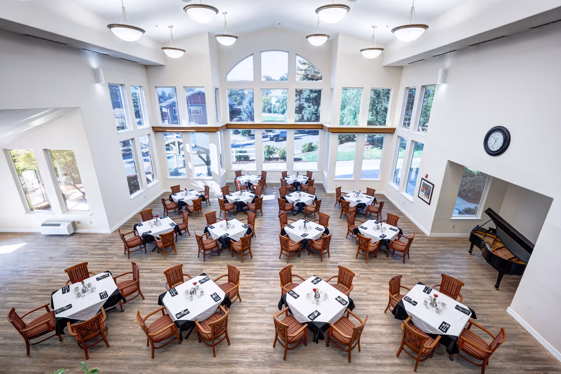 A spacious dining room with multiple round tables covered with white tablecloths and black overlays, each surrounded by wooden chairs with striped cushions. The room features large windows allowing natural light to fill the space, a grand piano in the corner, and a clock on the wall. The floor is wood, and the ceiling has several modern light fixtures.