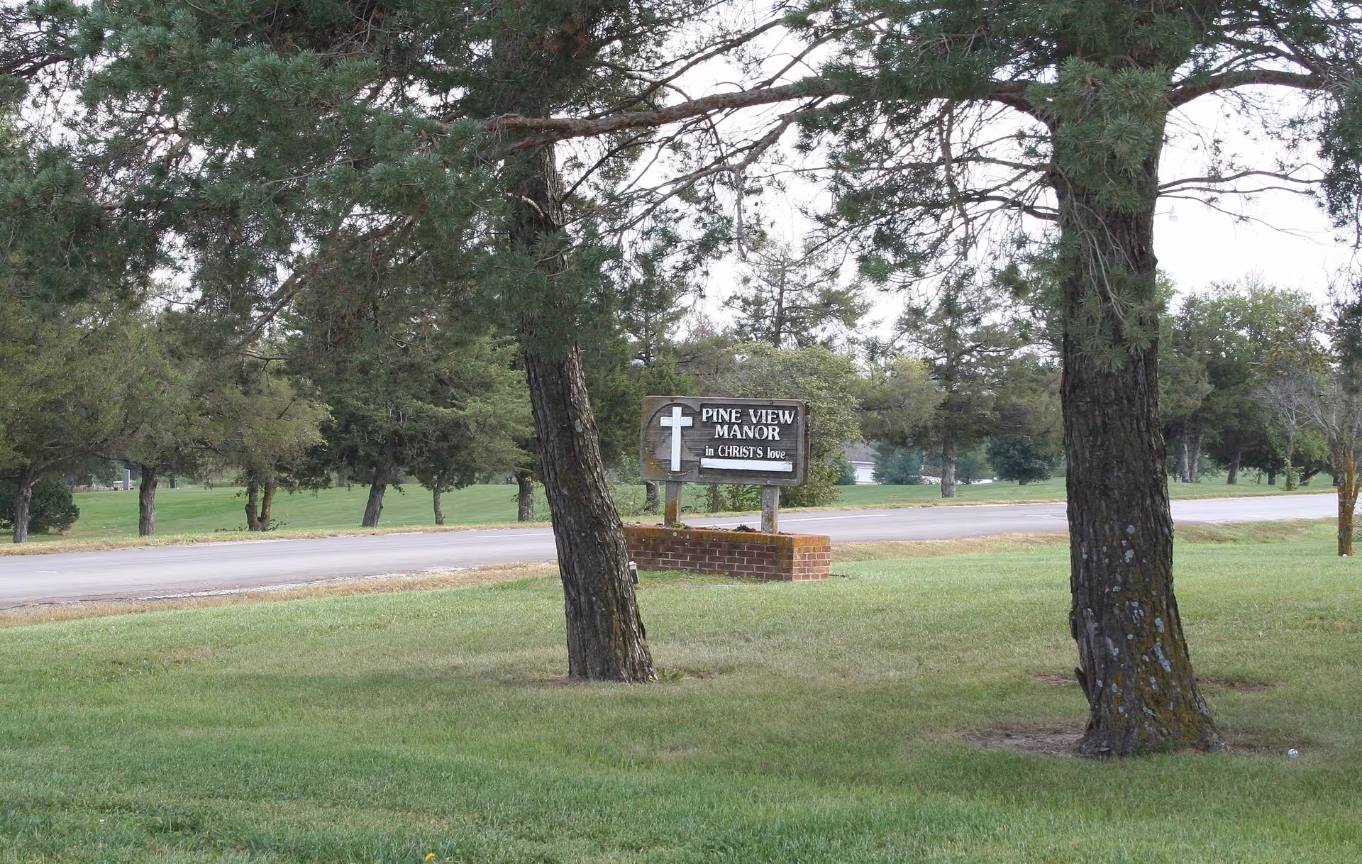 Grassy lawn and trees by a road with a brick-mounted sign reading 'Pine View Manor'.