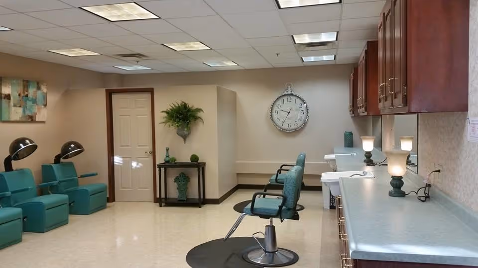 Interior view of a salon area in a senior living facility with teal salon chairs, hair drying stations, a large wall clock, wooden cabinets, and a countertop with lamps and sinks.