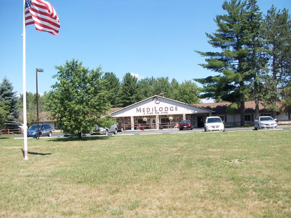 Front exterior of the Medilodge building with its entrance sign, an American flag on a pole, and several parked cars on a grassy lawn.