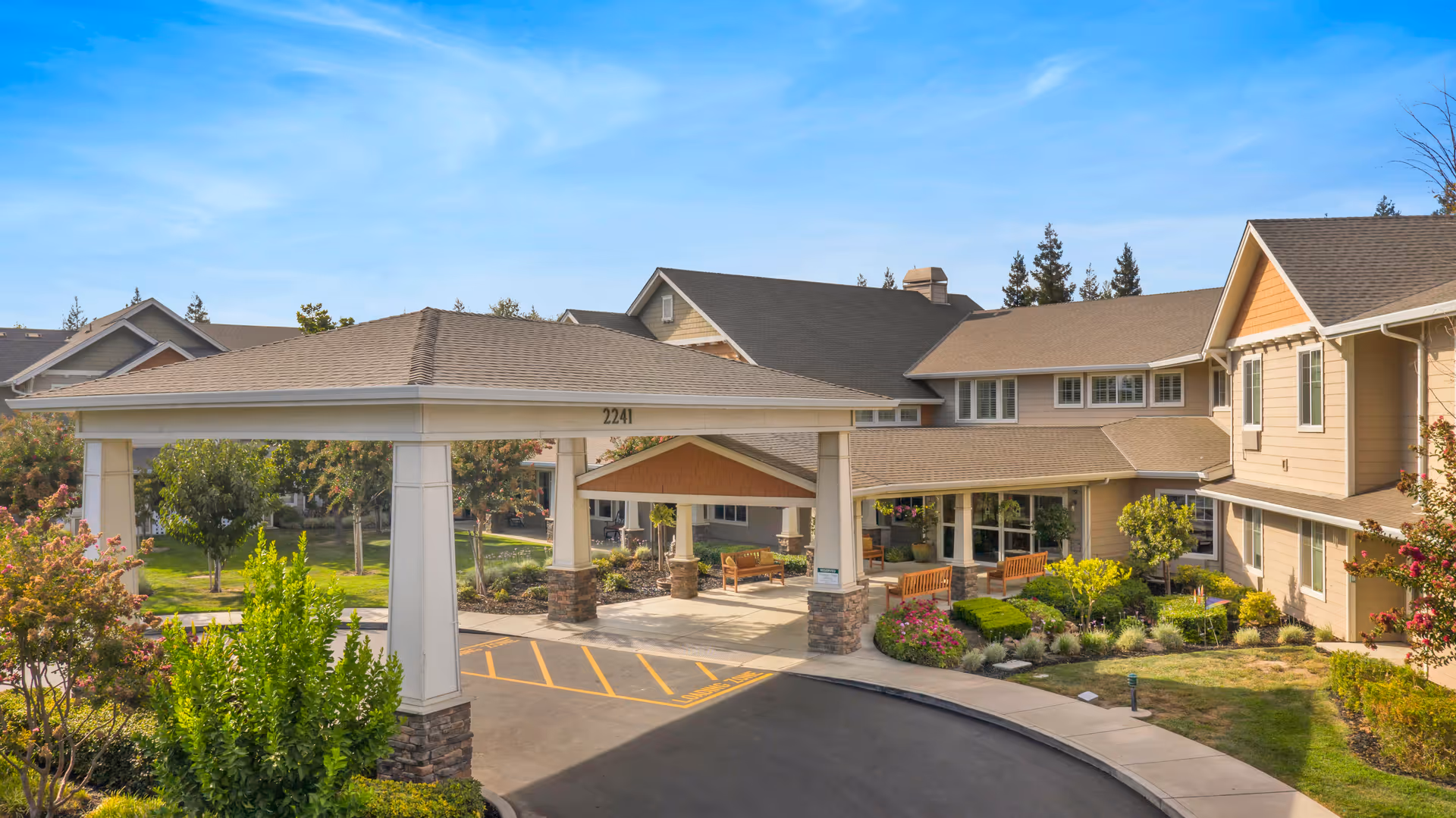 Exterior view of The Commons at Union Ranch senior living facility showing the main entrance with a covered drop-off area, landscaped gardens, benches, and a curved driveway under a clear blue sky.