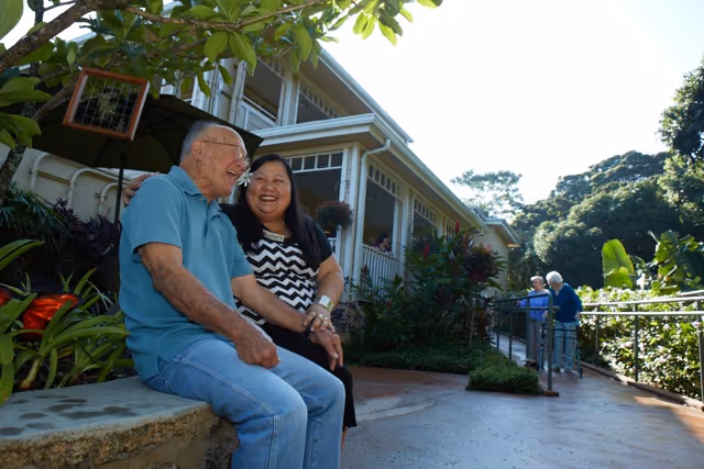 An elderly man and a woman sitting on a stone bench outside a residential building, smiling and enjoying each other's company. In the background, two other elderly people are walking along a pathway surrounded by lush greenery and plants under a clear sky.