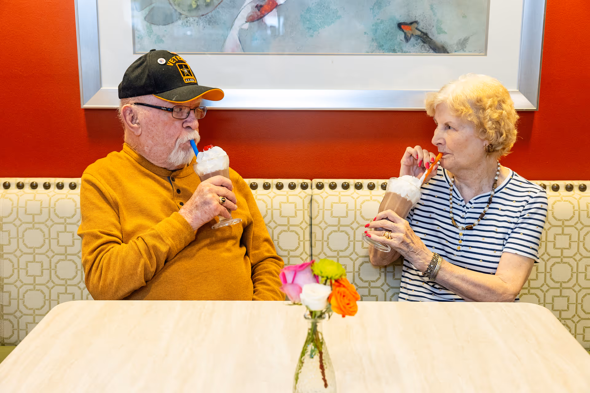 An elderly man and woman sitting at a table in a dining area, each drinking a milkshake topped with whipped cream. The man is wearing a black cap and mustard yellow sweater, while the woman is in a striped shirt. A small vase with colorful flowers is on the table in front of them, and a framed artwork hangs on the red wall behind them.