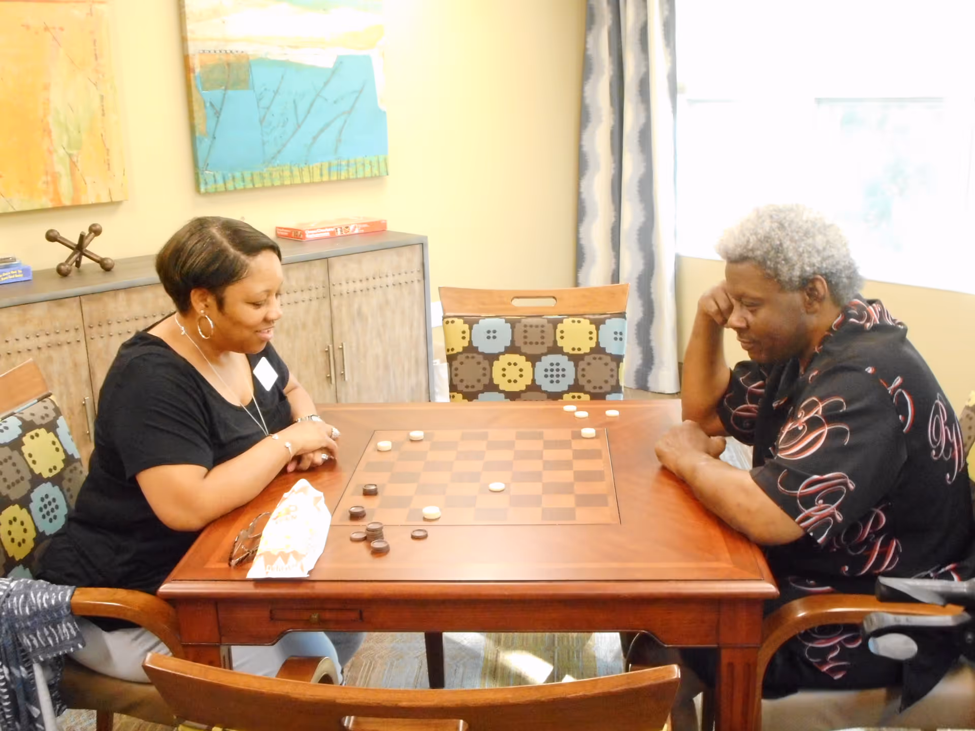 Two women sitting at a wooden table playing a game of checkers in a well-lit room with patterned chairs and colorful artwork on the wall behind them.
