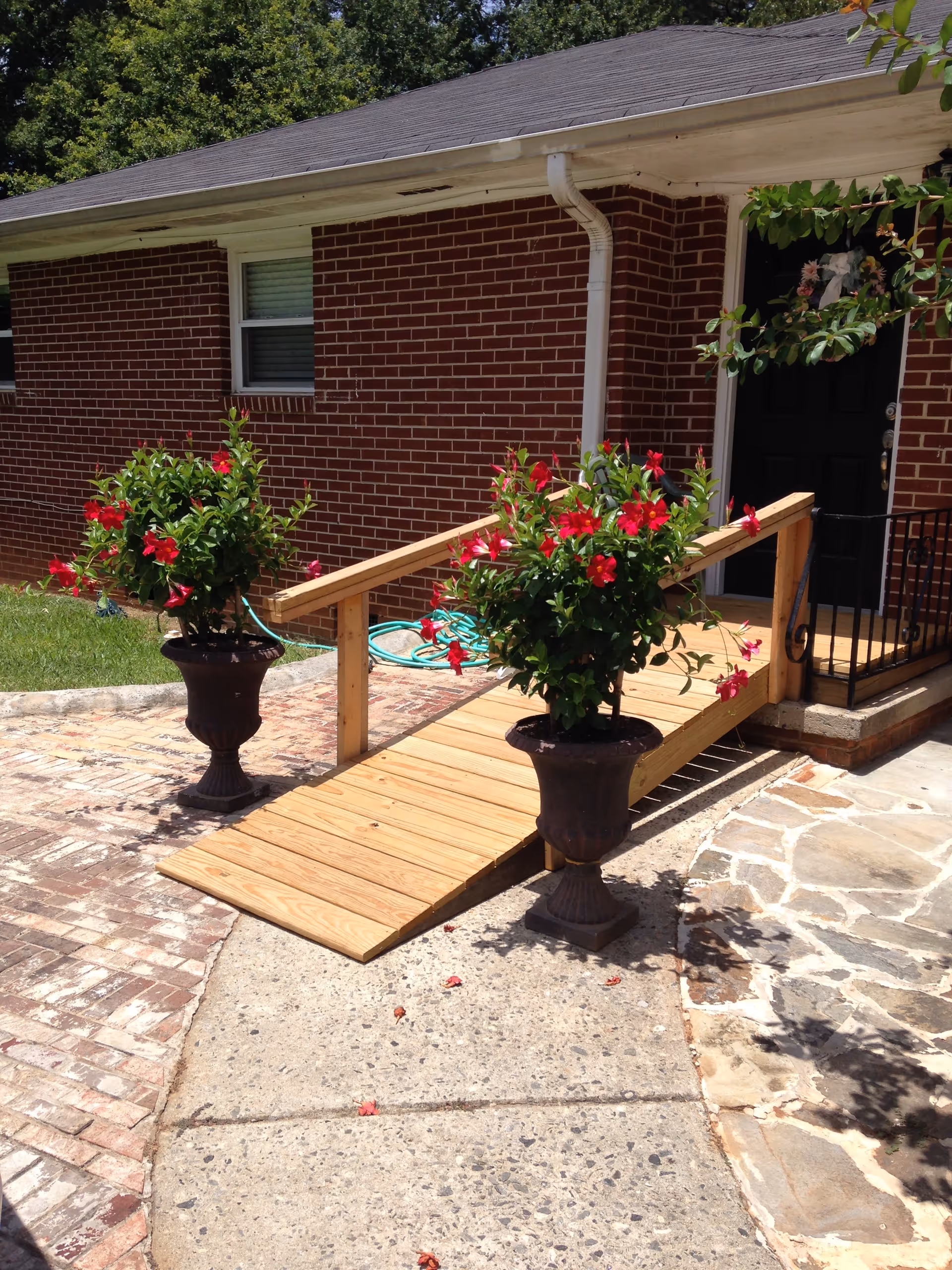 Exterior view of a brick building with a wooden wheelchair ramp leading to a black door decorated with a floral wreath. Two large planters with red flowers flank the ramp. The surrounding area includes a concrete walkway, brick pavement, and stone tiles.