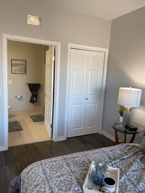 View of a bedroom with a patterned bedspread, a glass side table with a lamp and decorative items, and an open door leading to a bathroom with a towel hanging and a toilet paper holder visible.