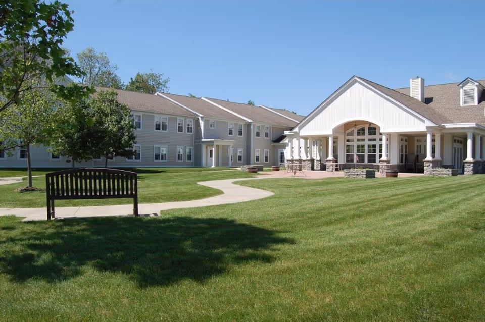Outdoor view of Yorktown Assisted Living Residence showing a well-maintained lawn with a wooden bench, a paved walkway, trees, and a large building with white siding and a covered porch under a clear blue sky.