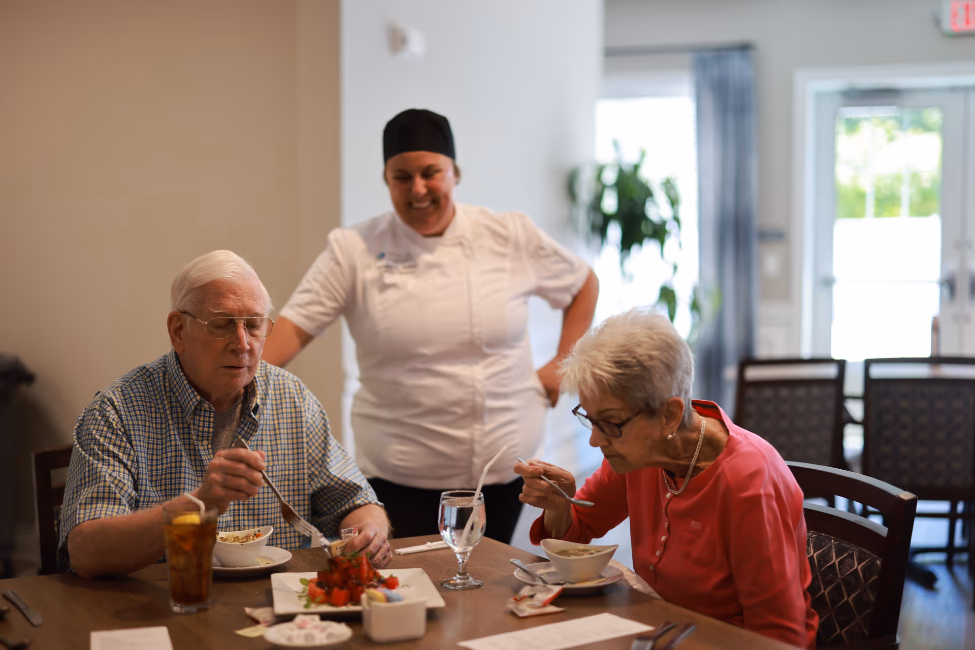 An elderly man and woman are seated at a dining table eating a meal. The man is wearing glasses and a checkered shirt, while the woman is wearing glasses and a pink top. A smiling chef in a white uniform and black hat stands behind them, observing. The dining area has chairs and tables, with a window and door in the background letting in natural light.