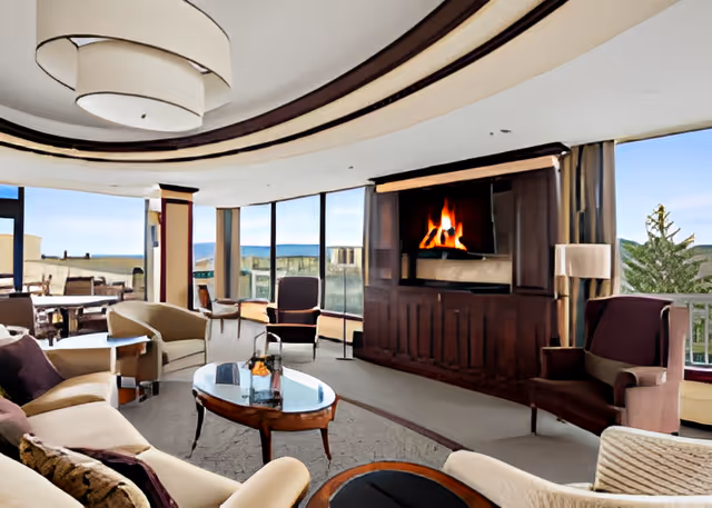 A spacious and elegant living room with large windows offering a scenic view. The room features a modern fireplace with a wooden mantel, comfortable armchairs, a sofa, a glass coffee table, and a round dining table in the background. The ceiling has a stylish circular light fixture.