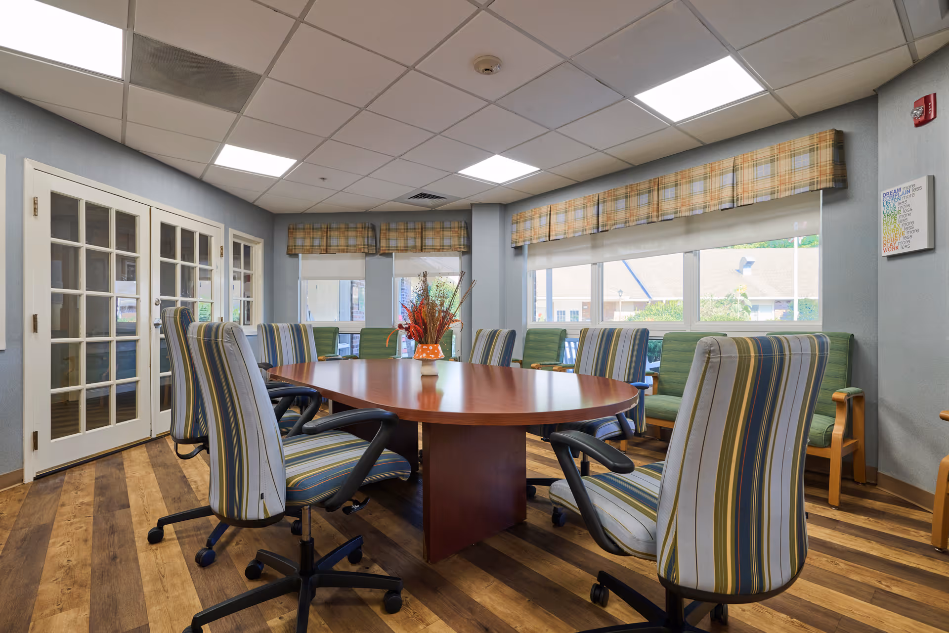 A conference room with a large oval wooden table surrounded by striped office chairs on wheels. The room has wood flooring, large windows with plaid valances, and a small green couch along the wall. French doors with glass panes are visible on the left side.