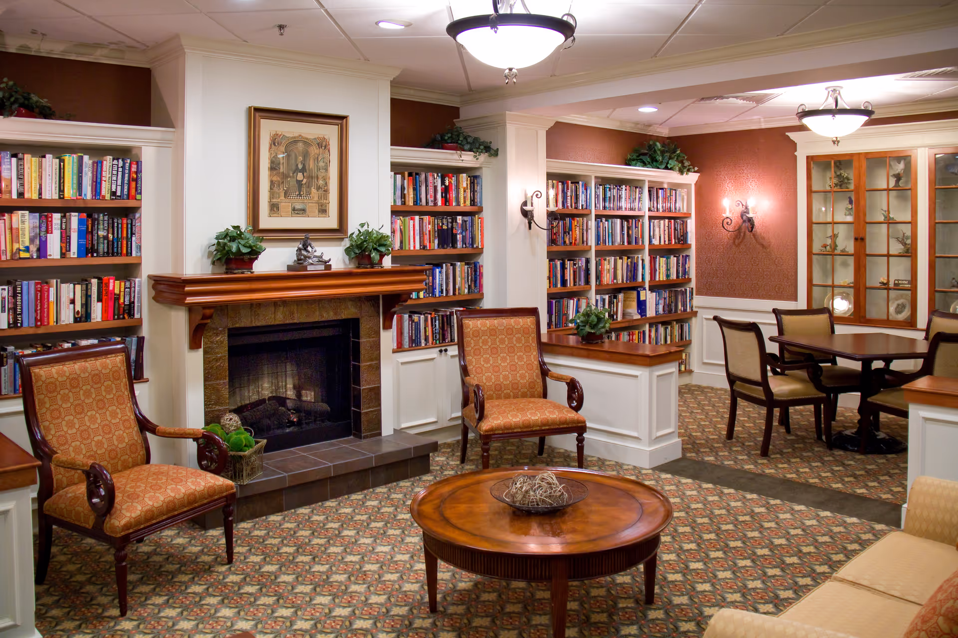 A cozy library room with built-in bookshelves filled with books, a fireplace with a wooden mantle, two upholstered armchairs, a round wooden coffee table with decorative items, and a dining area with a table and chairs. The room has warm lighting, patterned carpet, and decorative plants on shelves and mantle.