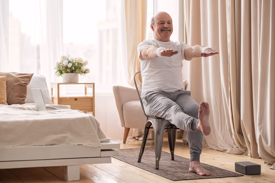 An elderly man sitting on a metal chair in a bedroom, performing a seated leg exercise with his arms extended forward. The room has a bed with pillows, a small wooden side table with a vase of flowers, a beige armchair, and large windows with sheer curtains allowing natural light to fill the space.