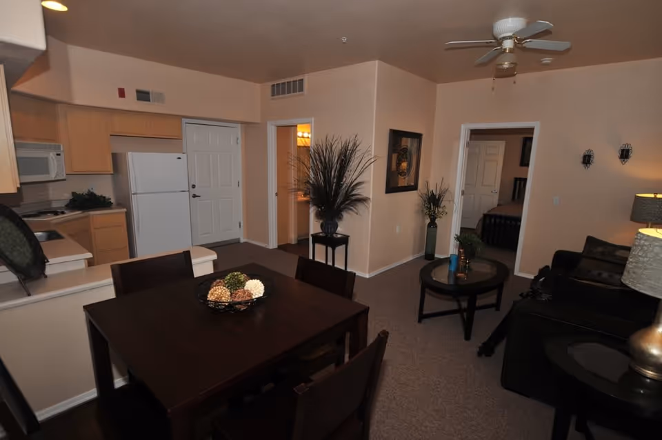 Interior view of a retirement home apartment showing a dining area with a dark wooden table and chairs, a kitchen with light wood cabinets, a white refrigerator and microwave, a living area with a dark sofa, coffee table, and side table with a lamp, beige walls, carpeted floor, ceiling fan, and decorative plants and artwork on the walls.
