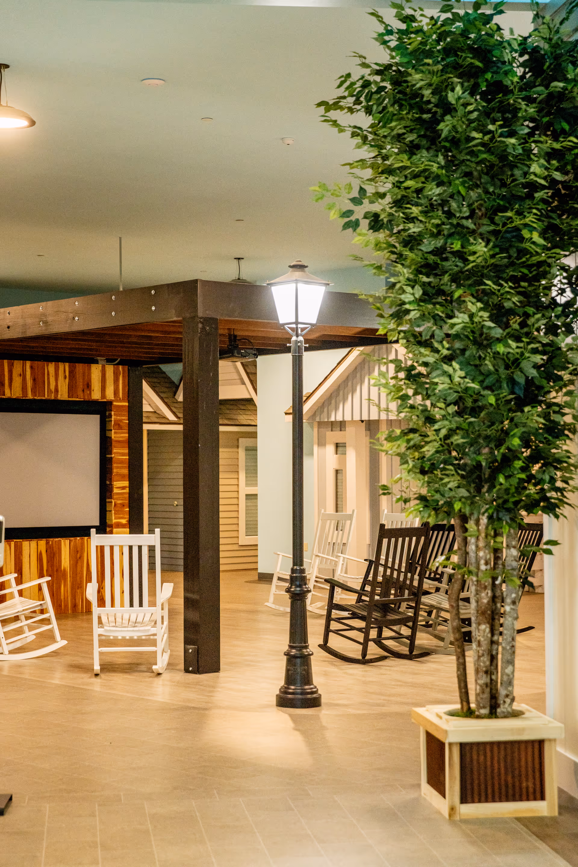 An indoor common area featuring white and black rocking chairs, a lamppost-style light, and a large potted tree.