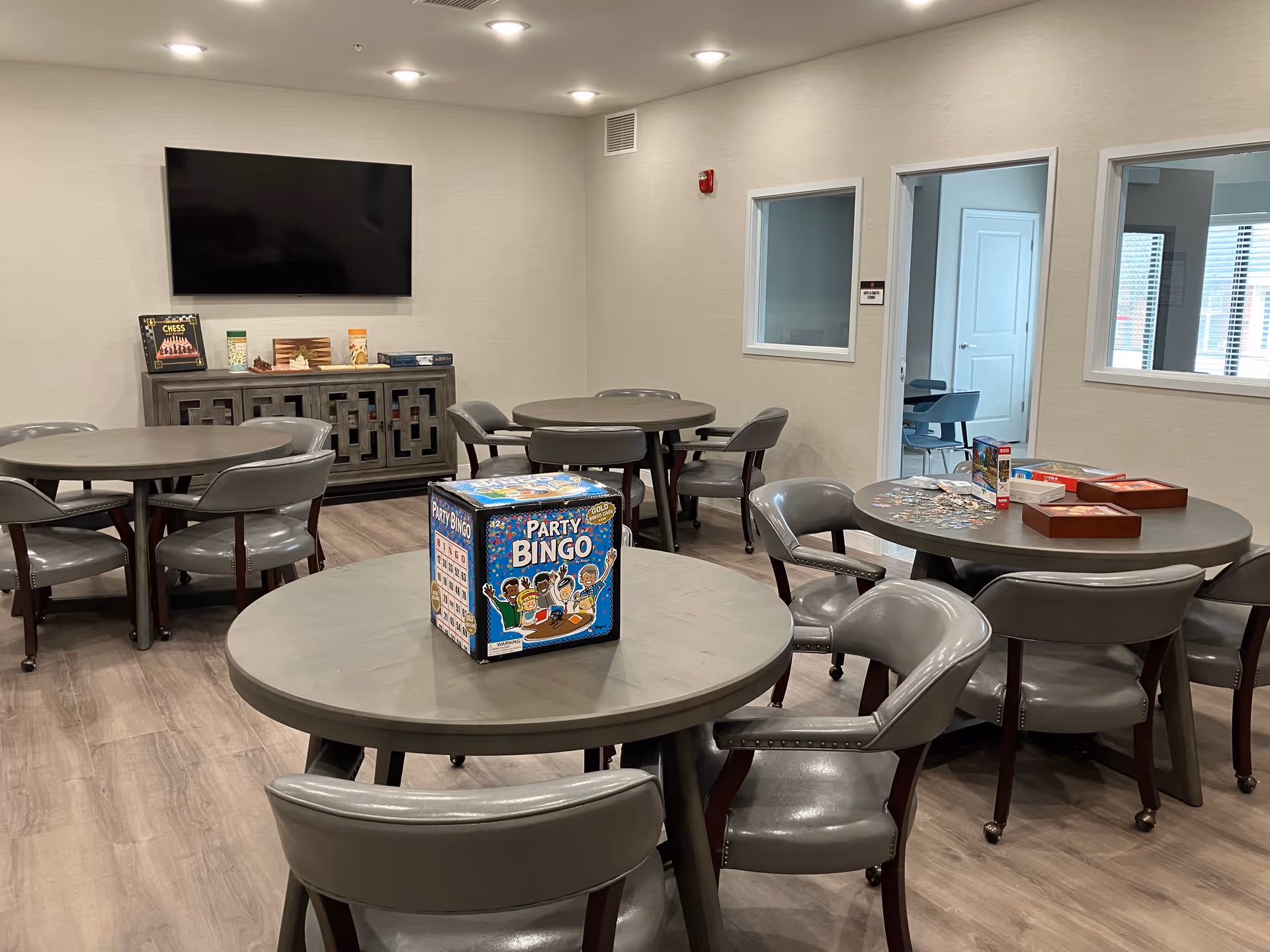 A community room with several round tables and gray chairs arranged around them. On the tables are board games and puzzles, including a Party Bingo game and a partially completed jigsaw puzzle. A large flat-screen TV is mounted on the wall above a cabinet that holds more games. The room has light-colored walls and wood flooring, with windows and a door leading to another room.