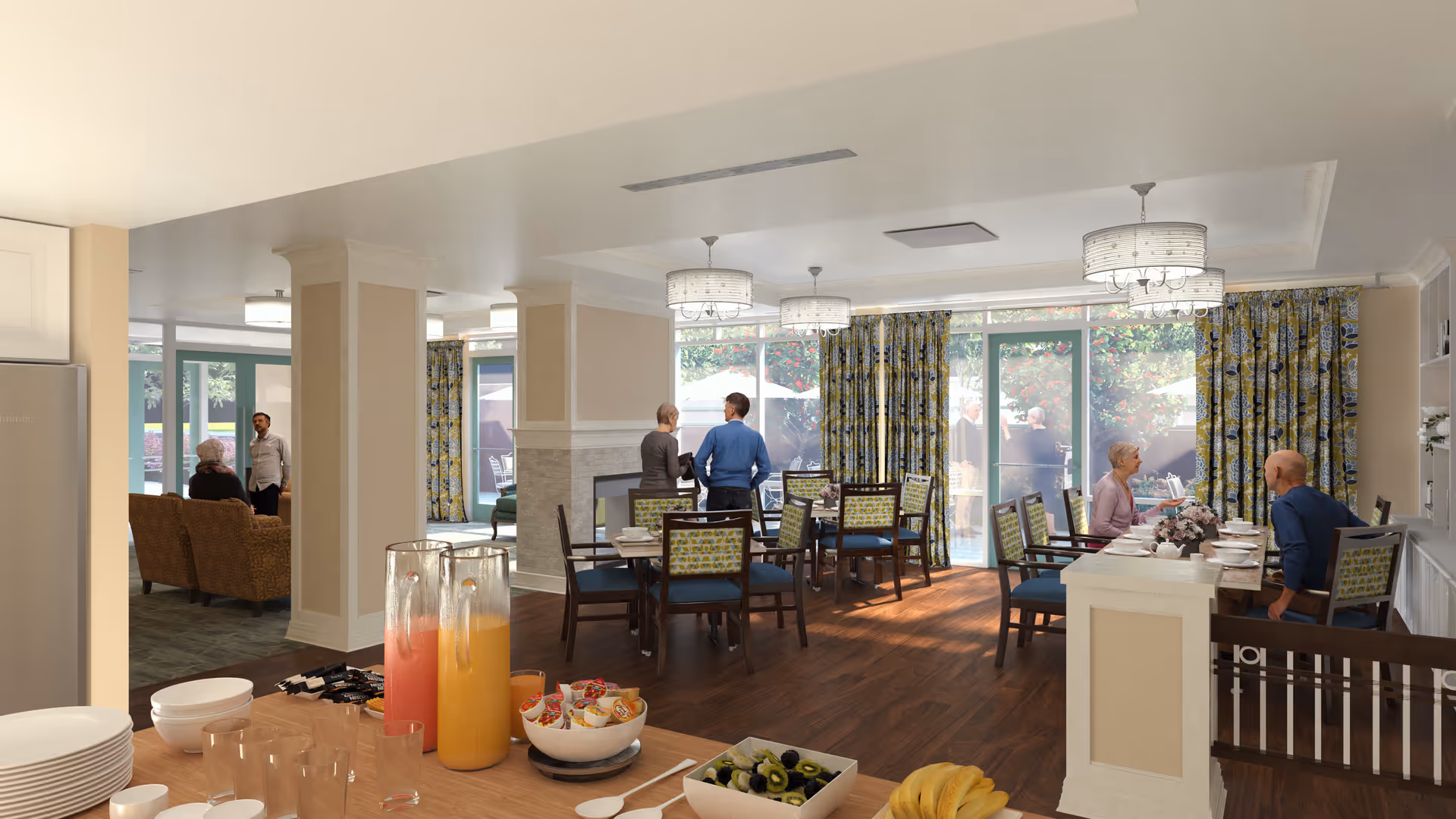 A bright dining room in a senior living facility with several tables and chairs. Two large glass pitchers filled with orange and pink beverages sit on a counter along with bowls of fruit and snacks. Several seniors and staff members are seen sitting and standing, engaging in conversation. Large windows with patterned curtains allow natural light to fill the room, and there is a cozy seating area visible in the background.