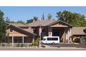 Exterior view of a single-story building with a beige facade and a sloped roof, featuring large windows and an entrance with a covered porch. A white shuttle van is parked in front of the building, and there are trees and shrubs around the property under a clear blue sky.