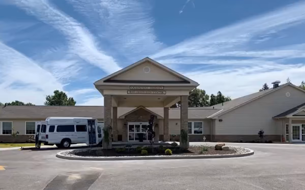Front exterior view of The Center for Rehabilitation at Hampton Woods building with a covered entrance, a white van parked on the left side, and a circular driveway with landscaping in front. The sky is blue with wispy clouds.