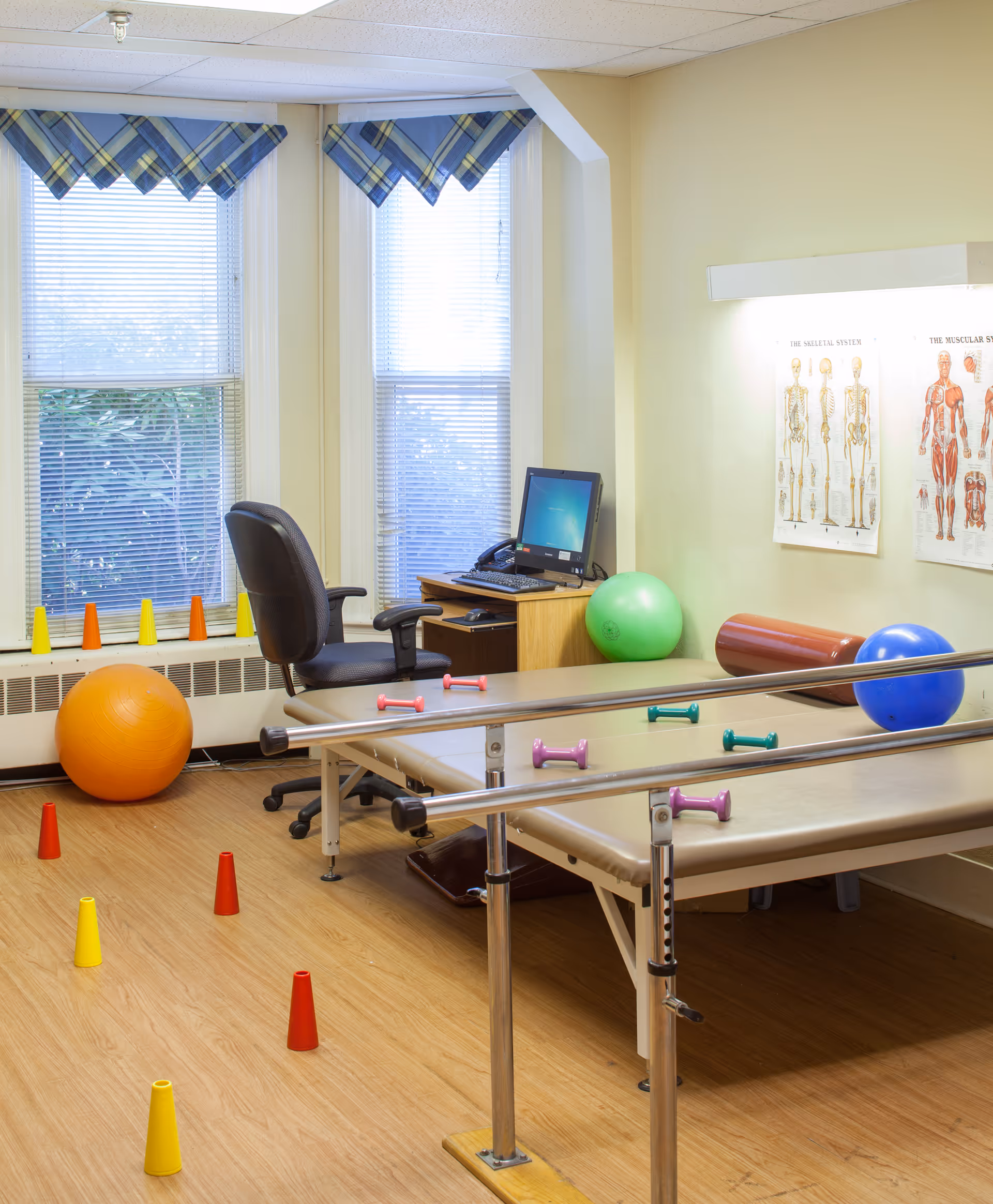 A physical therapy or rehabilitation room with parallel bars, exercise balls, small dumbbells, and colorful cones arranged on a wooden floor. There is a desk with a computer and an office chair near two large windows with blue plaid valances. Anatomical charts of the muscular and skeletal systems are displayed on the wall.