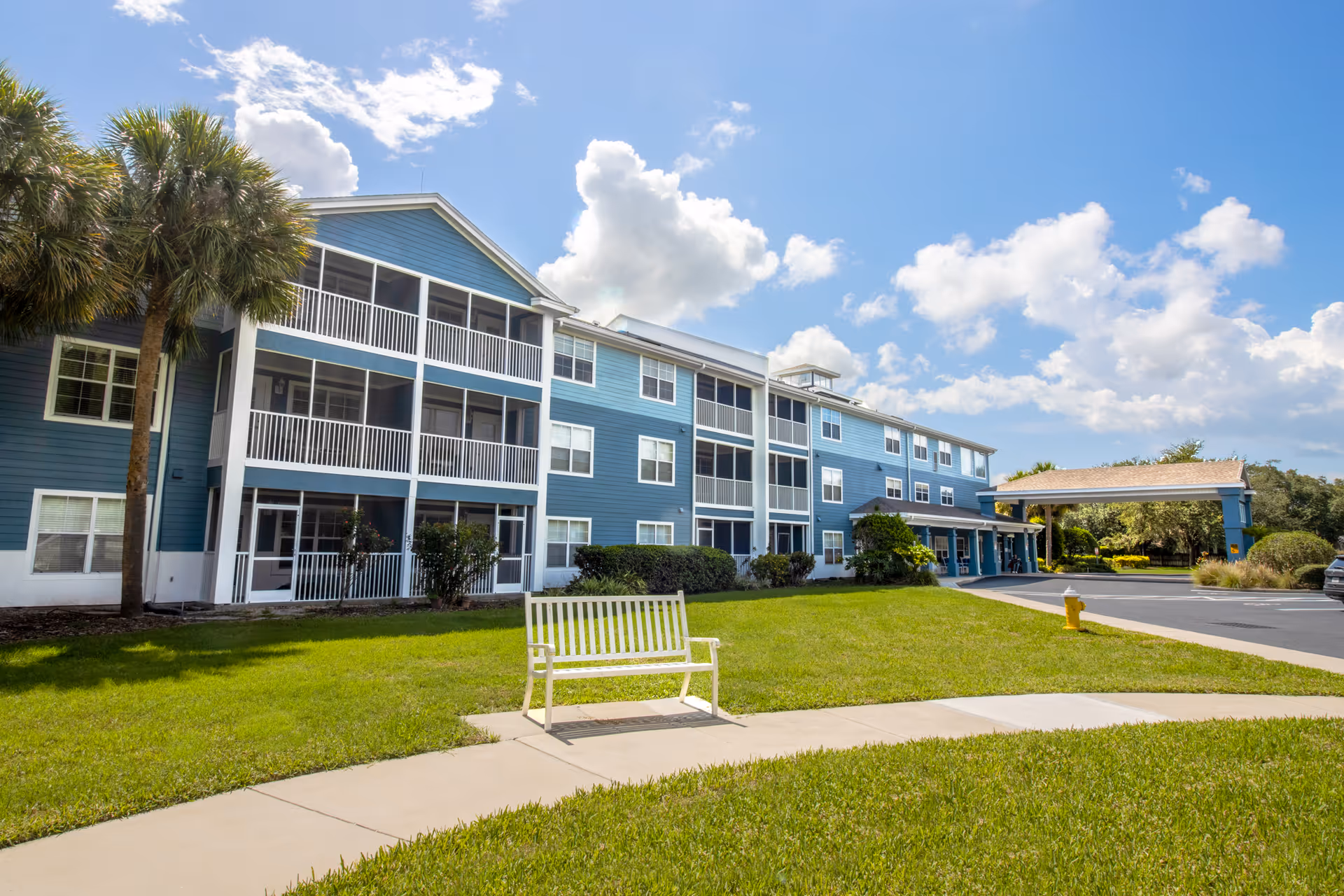 Front exterior of a blue three-story senior living building with screened balconies, a bench on the lawn, and a covered entrance.
