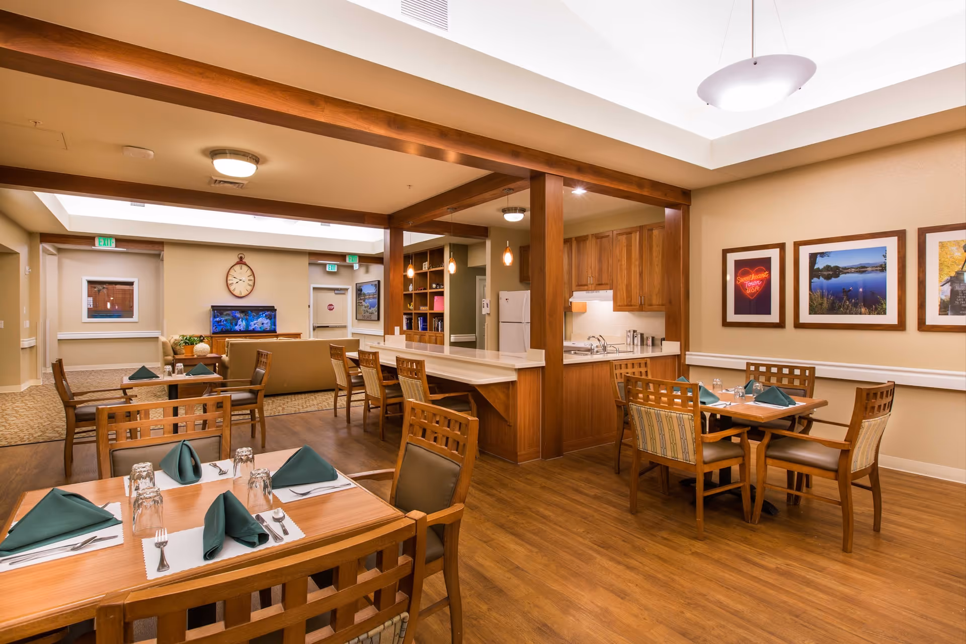 A dining area in a senior living facility with wooden tables and chairs set with green napkins and glassware. The room features warm lighting, wood flooring, and a small kitchen area with wooden cabinets and a white refrigerator. In the background, there is a seating area with a couch, a wall clock, and a fish tank. The walls are decorated with framed pictures.