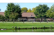 Single-story building with a porch behind a fenced grassy lawn and a pond with several geese, backed by trees.