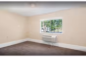 Empty room with beige walls, a window showing green trees outside, a brown carpeted floor, and a wall-mounted air conditioning unit below the window.