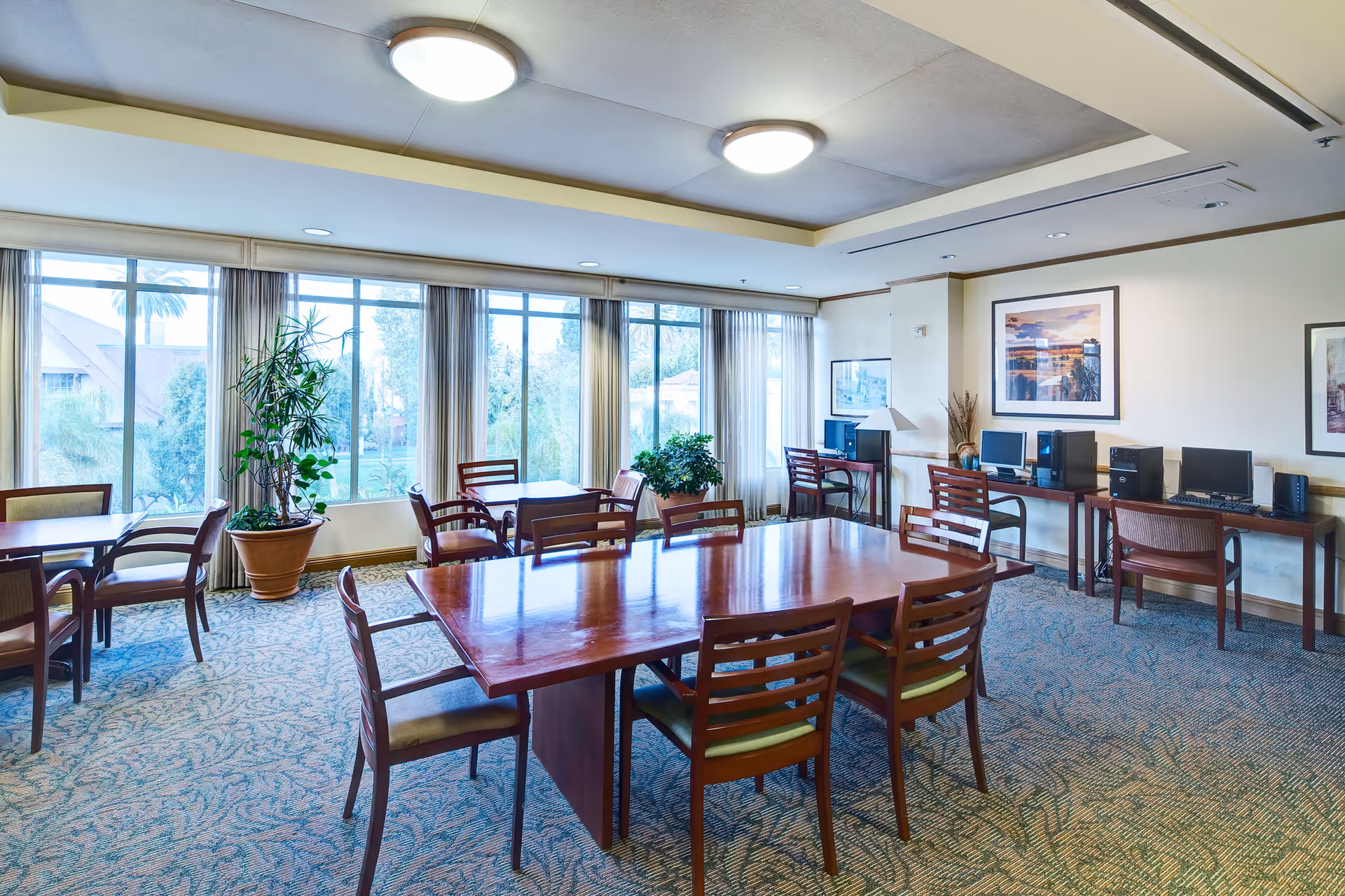 A bright communal dining/activity room with wooden tables and chairs, large windows, potted plants, and computer stations along one wall.
