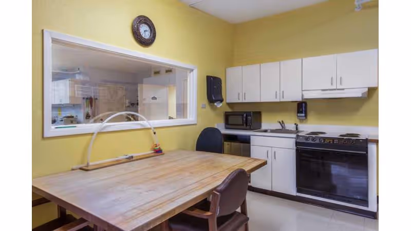 A small kitchen area with yellow walls, white cabinets, a microwave, a stove, and a wooden table with chairs. There is a large window on one wall showing another room, and a clock above the window.